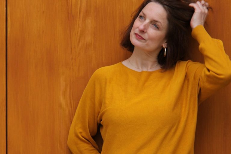 Frau mit braunen Haaren in gelbem Oberteil vor einer Holzoberfläche.